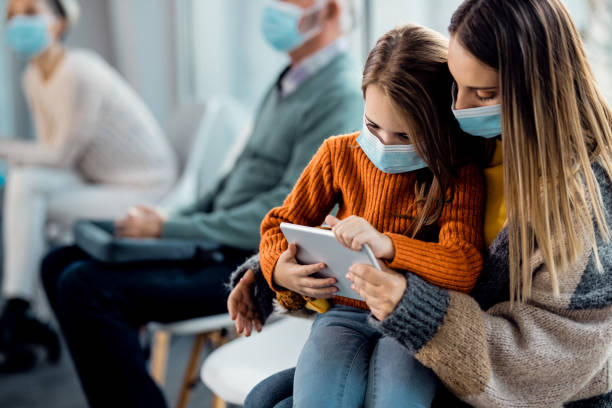 Mother and daughter with face masks using touchpad while waiting at medical clinic. Small girl and her mother wearing protective face masks and surfing the net while sitting in a waiting room at the hospital. waiting room stock pictures, royalty-free photos & images