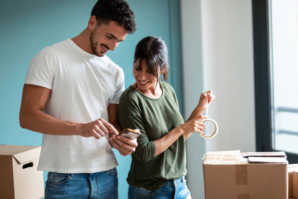 Attractive young couple packing boxes to move to new flat while looking mobile phone in living room at home.