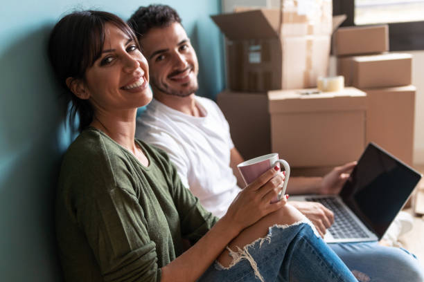 Beautiful young couple searching ideas with laptop while drinking a cup of coffee looking at camera sitting on the floor.