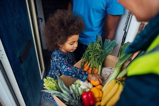 Over the shoulder view of a mature woman delivering a box full of fresh vegetables to an unrecognizable mid-adult male and his daughter.