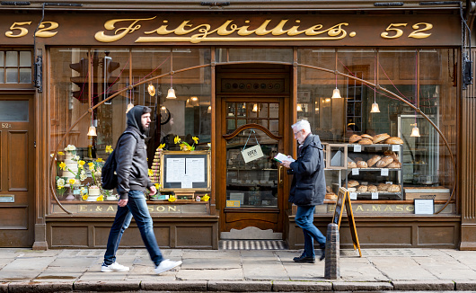 People sit inside the well-known Fitzbillies, a traditional bakery and café on Trumpington Street in Cambridge, England. There is a man walking past reading a book
