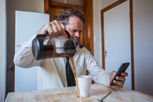 A working male accidently focuses on his phone rather than the coffee he is making, he spills coffee all over the kitchen table at the office.