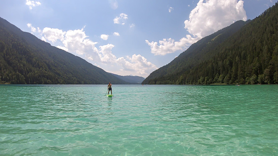 A man stand-up paddling on the crystal clear water of Weissensee lake in Austrian Alps. The lake is surrounded by high mountains. Exercising in the nature. Serenity and calmness.