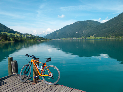 A bike parked on a wooden pier by the Weissensee lake in Austria. The lake is surrounded by the high Alps. Exercising on the free air. Many mountain chains in the back. Sunny day. Exploration
