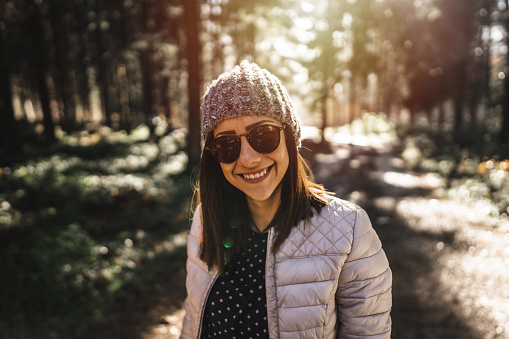 Portrait of beautiful young woman with sunglasses and cap in forest on sunny day, smiling and looking at camera
