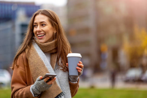 istock Smiling young woman with smart phone and coffee cup outdoors at urban setting 1286734285