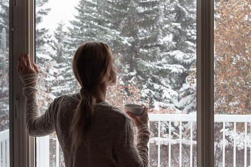 Woman standing in living room in home holding a coffee cup in one hand and looking out the window to the snow covered trees in winter, wearing tan sweater