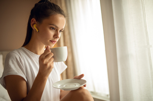 Beautiful young woman in a white T-shirt, sitting on the bed in her bedroom with headphones and enjoying her morning coffee in music