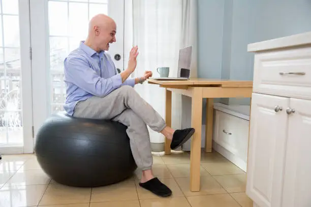 Man wears a dress shirt and jogging pants and sits on a yoga ball while on a business work from home zoom call on the laptop