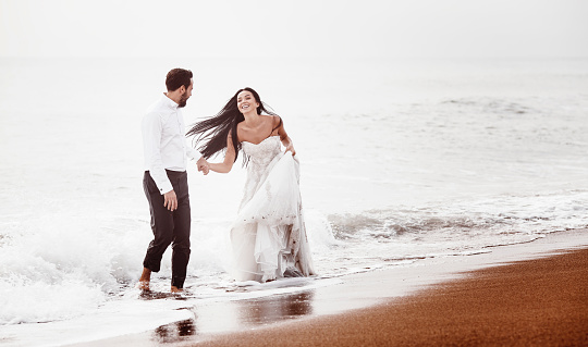 Young bride and groom happily run by the sea in their wedding clothes.