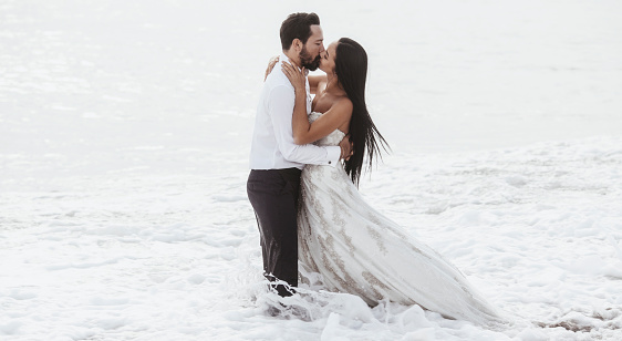 Young bride and groom kissing each other in the waves by the sea.