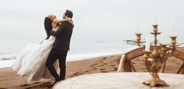 Young couple celebrate on the beach after their wedding ceremony. The bride is happily hugging the groom.