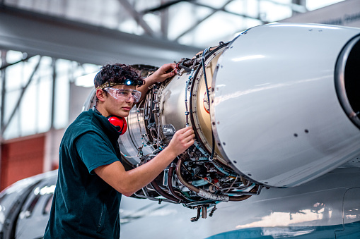 Young male aircraft maintenance mechanic in the airplane hangar repairing a jet engine properly equipped.