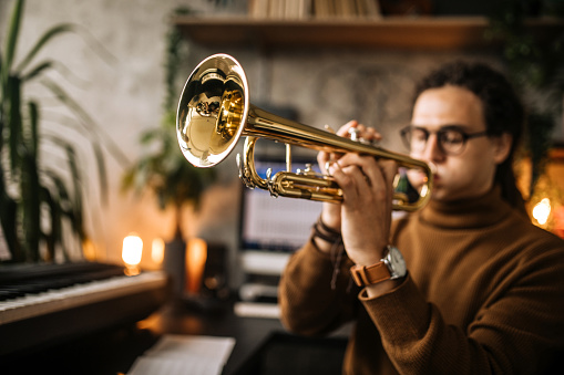 Young handsome musician practicing trumpet playing in his home musical studio.