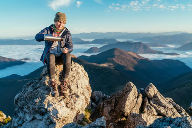 istock Active backpacker hiker woman sitting on the mountain summit cliff, pouring a tea from a thermos flask and enjoying mountains valley covered with clouds view. Successful summit climbing concept image. 1286762797