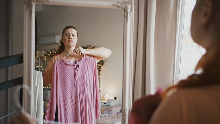 Kobieta trzyma przed lustrem różową sukienkę i rozważa ją jako okazję do chrztu.