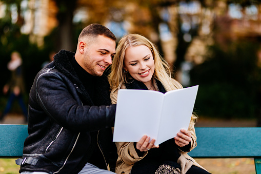 A young couple sits on a park bench and reads an empty magazine (mock-up) together.