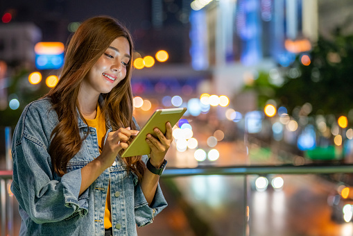 Happy young beautiful Asian woman using digital tablet in the city at night