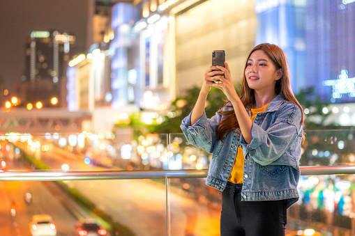 Happy Asian woman using smartphone taking a photo or recording video of city street at night