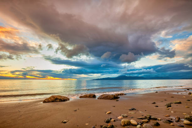 A beach during sunset. There are darker blue and orange clouds in the blue sky. The sea water moves quickly and it reflects the sky and the clouds.