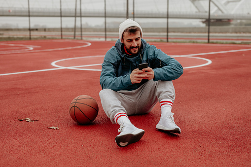Handsome cool sportsman in a jacket on the basketball court outdoors sitting and texting.
