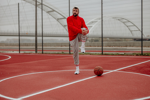 Fit bearded male in a red sweatshirt warming up on a basketball court outdoors.