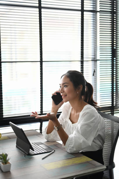 istock A cheerful young businesswoman is talking on mobile phone and using tablet while sitting at her working place. 1286801807