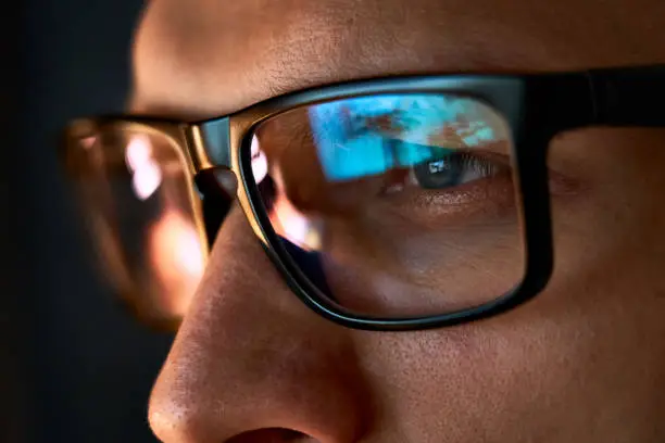 Photo of Close up view of focused businessman wears computer glasses for reducing eye strain blurred vision looking at pc screen with computer reflection using internet, reading, watching, working online late.