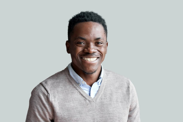 sorrindo alegre jovem adulto afro-americano homem de etnia olhando para a câmera em pé no fundo do home office. feliz e confiante negro posando para o rosto da frente de perto retrato. - foto de cabeça - fotografias e filmes do acervo