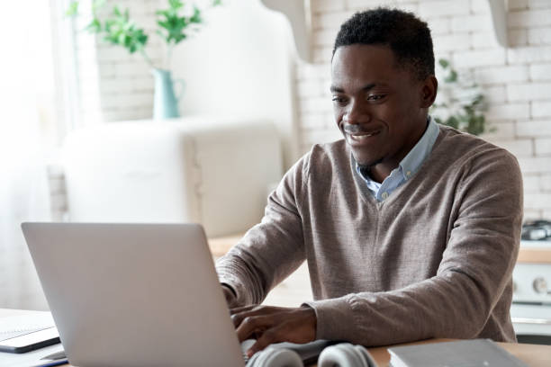 istock Smiling professional black business man using laptop computer working from home office. Happy african male customer makes ecommerce delivery order from supermarket typing on pc sits at kitchen table. 1286810740