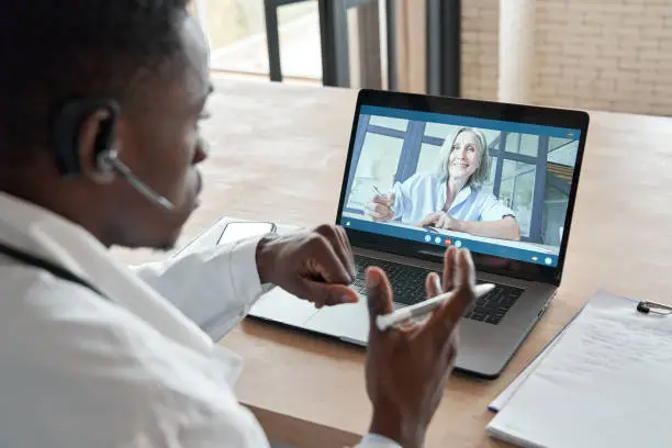Photo of Black male doctor consulting senior old patient by telemedicine online video call. African physician using telehealth medical chat virtual healthcare appointment on laptop computer. Over shoulder view
