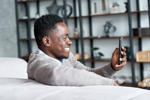 istock Smiling african american man holding smart phone using social media apps sitting on couch at home. Black guy texting, remote learning, ordering distance delivery or chatting online in application. 1286810749