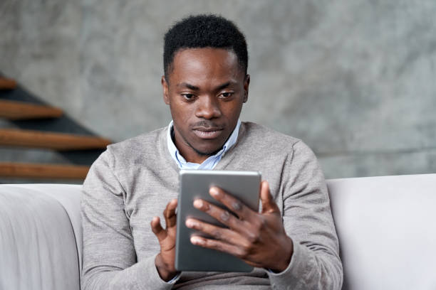 istock Young adult smart african american ethnic man holding digital tablet using ecommerce apps on tab computer sitting on couch at home for reading, checking email, ordering online or browsing internet. 1286810751