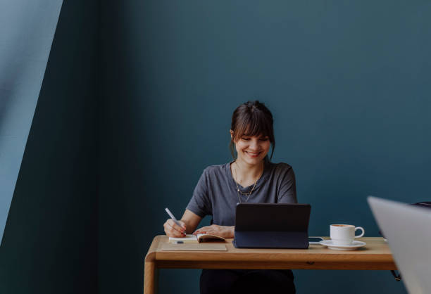 Women in Business: a Smiling Young Businesswoman Using a Digital Tablet at her Office Happy young woman, casually dressed, using technology at work (blue background, copy space). freelance work stock pictures, royalty-free photos & images