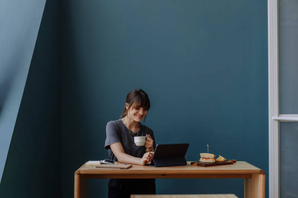 Women in Business: a Smiling Young Businesswoman Using a Digital Tablet and Holding a Cup of Coffee at her Office Happy young woman, casually dressed, using technology at work (blue background, copy space). standing desk photos stock pictures, royalty-free photos & images
