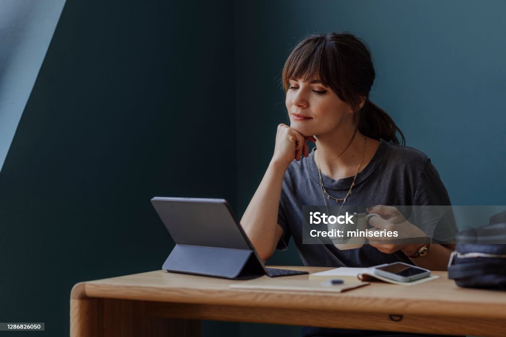 Women in Business: a Young Businesswoman Using a Digital Tablet and Holding a Cup of Coffee at her Office Happy young woman, casually dressed, using technology at work (blue background, copy space). Digital Tablet Stock Photo