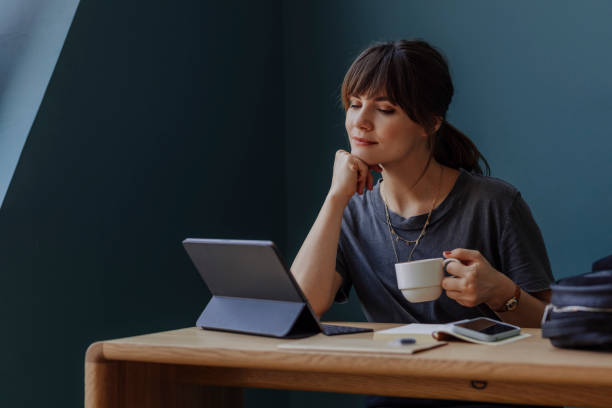 Women in Business: a Young Businesswoman Using a Digital Tablet and Holding a Cup of Coffee at her Office Happy young woman, casually dressed, using technology at work (blue background, copy space). standing desk photos stock pictures, royalty-free photos & images
