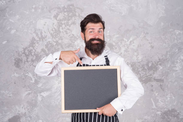 istock looking great. best cuisine here. welcome on board. brutal man hipster hold blackboard, copy space. coffee to go. nicest price in town. happy bearded chef pointing finger. successful cook career 1286836989