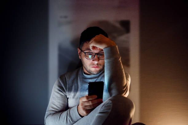 The social media addiction A 35-year-old guy is sitting in the dark room and using his smartphone. worried stock pictures, royalty-free photos & images