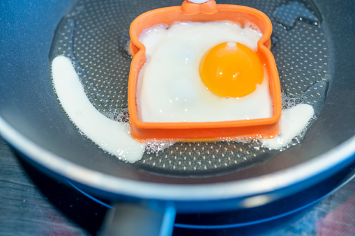 An egg frying in a non-stick teflon pan on an induction hob in a modern kitchen, it has been cooked in a mould shaped like a slice of bread.
