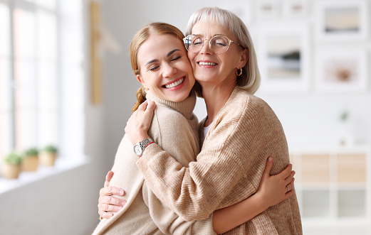 Delighted adult and senior women smiling with closed eyes and hugging each other on weekend day at home