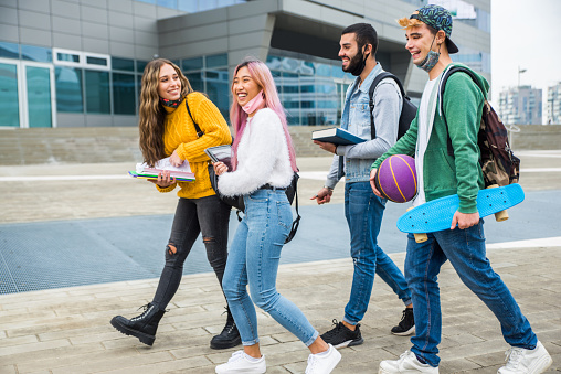 A small group of University students are seen walking outside on campus as they make their way to class in the morning.  They are dressed casually and have backpacks, shoulder bags and books in their hands.