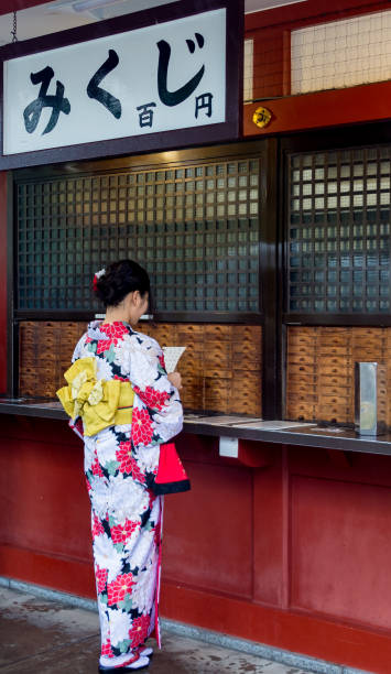 Girl in kimono reading Omikuji in Senso-ji Buddhist temple in Tokyo Japan Tokyo / Japan - October 19, 2017: Ancient Senso-ji Buddhist temple in Tokyo. Girl in kimono is reading Omikuji, Japanese fortune-telling paper strips that can be found at shrines and temples. sensoji stock pictures, royalty-free photos & images