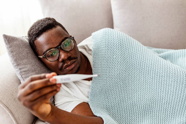 It can be worrying to see a temperature spike up Sick African-American man laying on sofa checking fever with thermometer. fever stock pictures, royalty-free photos & images