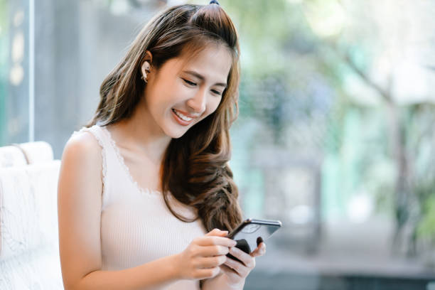 Portrait of smiling young Asian woman in trendy clothes listening to music or watching video via mobile phone with earbuds while sitting resting in cafe.Music Lover,Weekend Relaxation Leisure Concept. retrato de una joven asiática sonriente con ropa de moda escuchando música o viendo videos a través del teléfono móvil con auriculares mientras está sentada descansando en el café. amante de la música, concepto de ocio de relajación de fin de sema