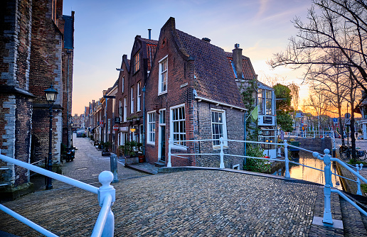 A see through an old street in the city of Delft. The sun is slowly setting and in the foreground is a bridge over a canal. The water reflects the surroundings.