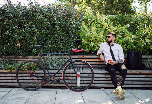 Portrait of young man commuter with bicycle sitting outdoors in city, using smartphone.