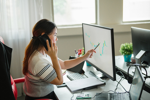 An Asian female Investment Banking Analyst talking on phone and accounting with statistics graphs on screen of computer at desktop.