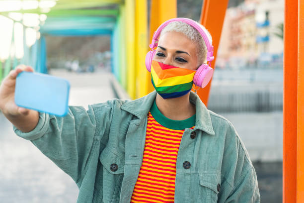 mujer joven que lleva máscara de orgullo gay escuchando música con auriculares inalámbricos y tomando selfie con teléfono inteligente móvil al aire libre - igualdad de género y concepto de tecnología - gay pride flag audio fotografías e imágenes de stock