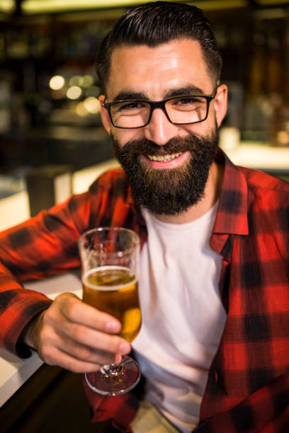 hipster em bar bebendo álcool. cerveja e tapas. posando sorrindo com luz ambiente. feche acima - beer glass mustache beer color image - fotografias e filmes do acervo